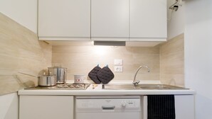 Cocina compacta con armarios blancos, lavavajillas, fregadero y encimera con tetera, olla y guantes de cocina.