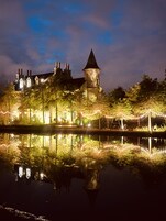 A historic building with a tower, surrounded by trees and a body of water reflecting the structure.