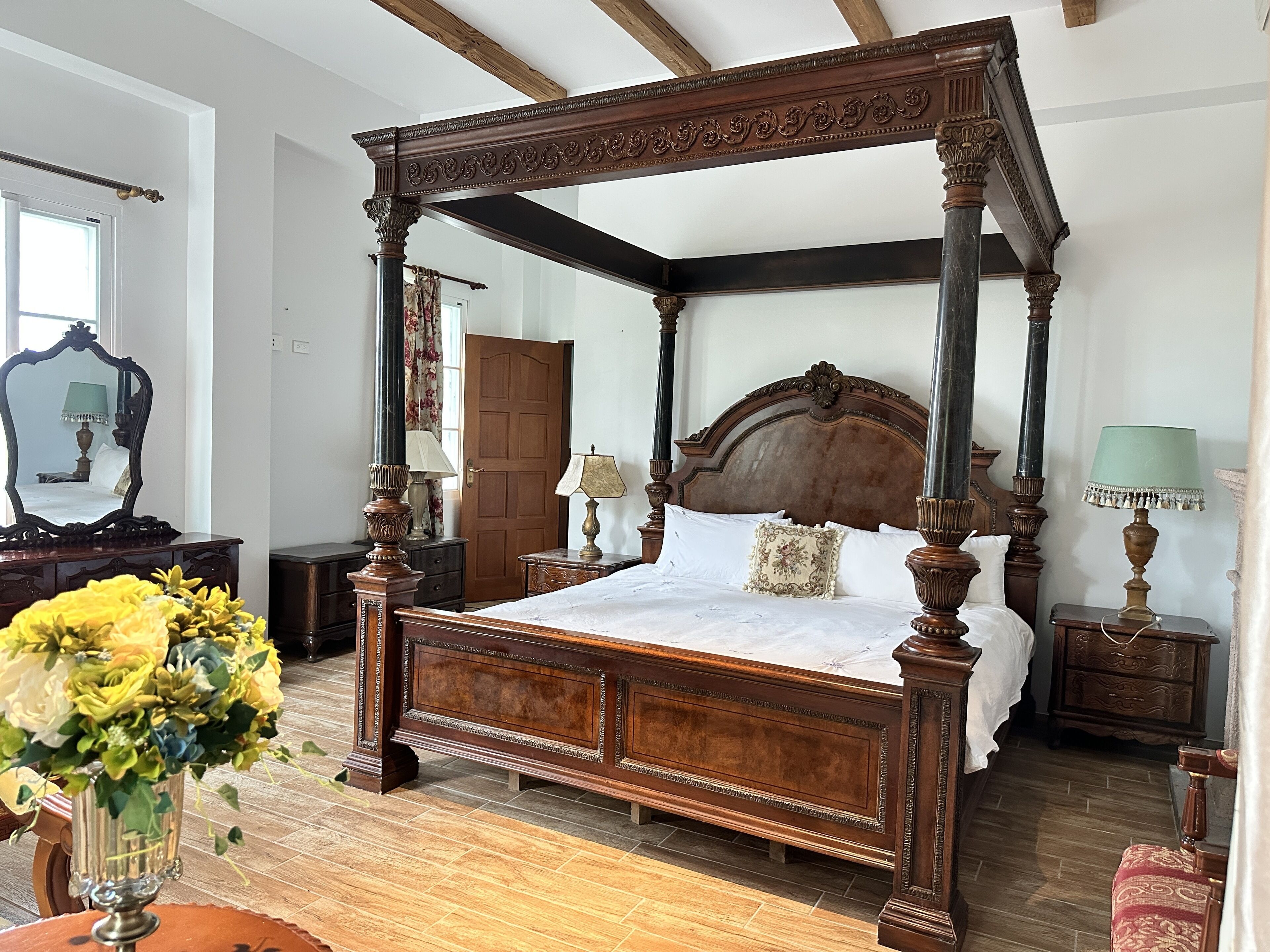 A four-poster bed with a wooden headboard, a vase of flowers, and a mirror on a dresser.
