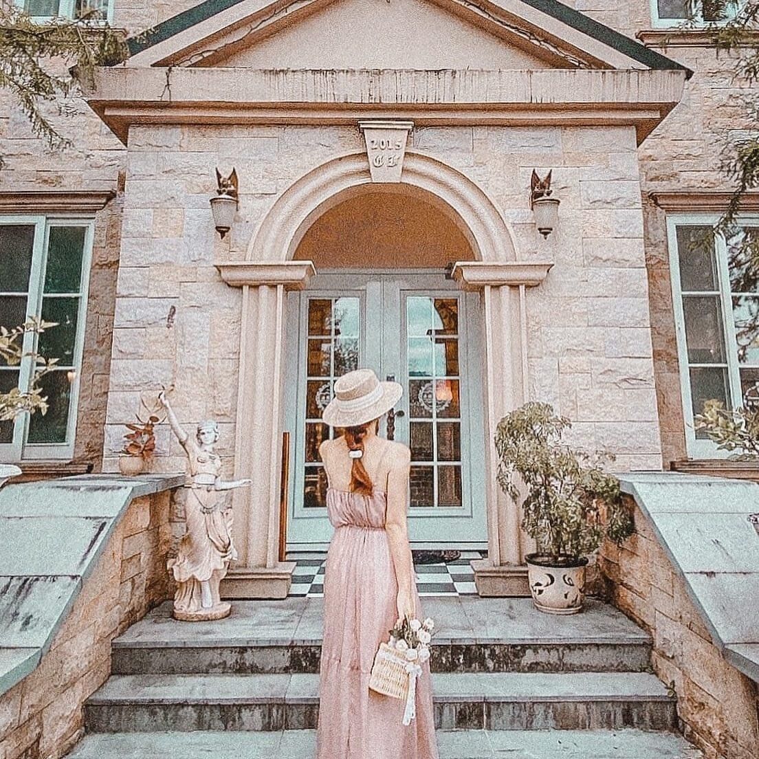 Una mujer con un vestido rosa está de pie en los escalones de un edificio de estilo arquitectónico clásico, con un arco central y columnas de piedra.