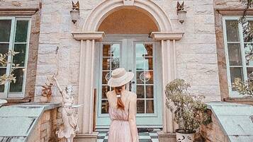 Una mujer con un vestido rosa está de pie en los escalones de un edificio de estilo arquitectónico clásico, con un arco central y columnas de piedra.