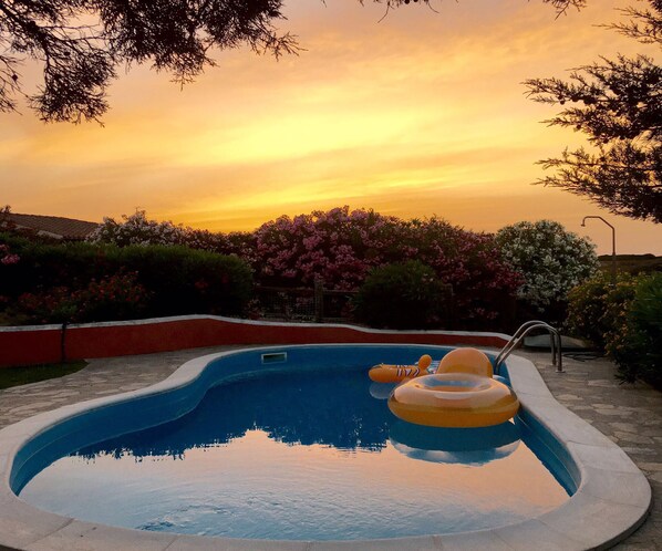 Piscina con vistas al atardecer, rodeada de arbustos florecientes y un muro rojo.
