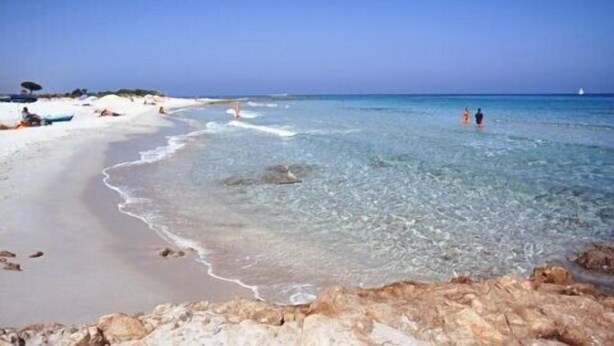 A beach with clear water, a sandy shore, and a rocky outcrop in the foreground.