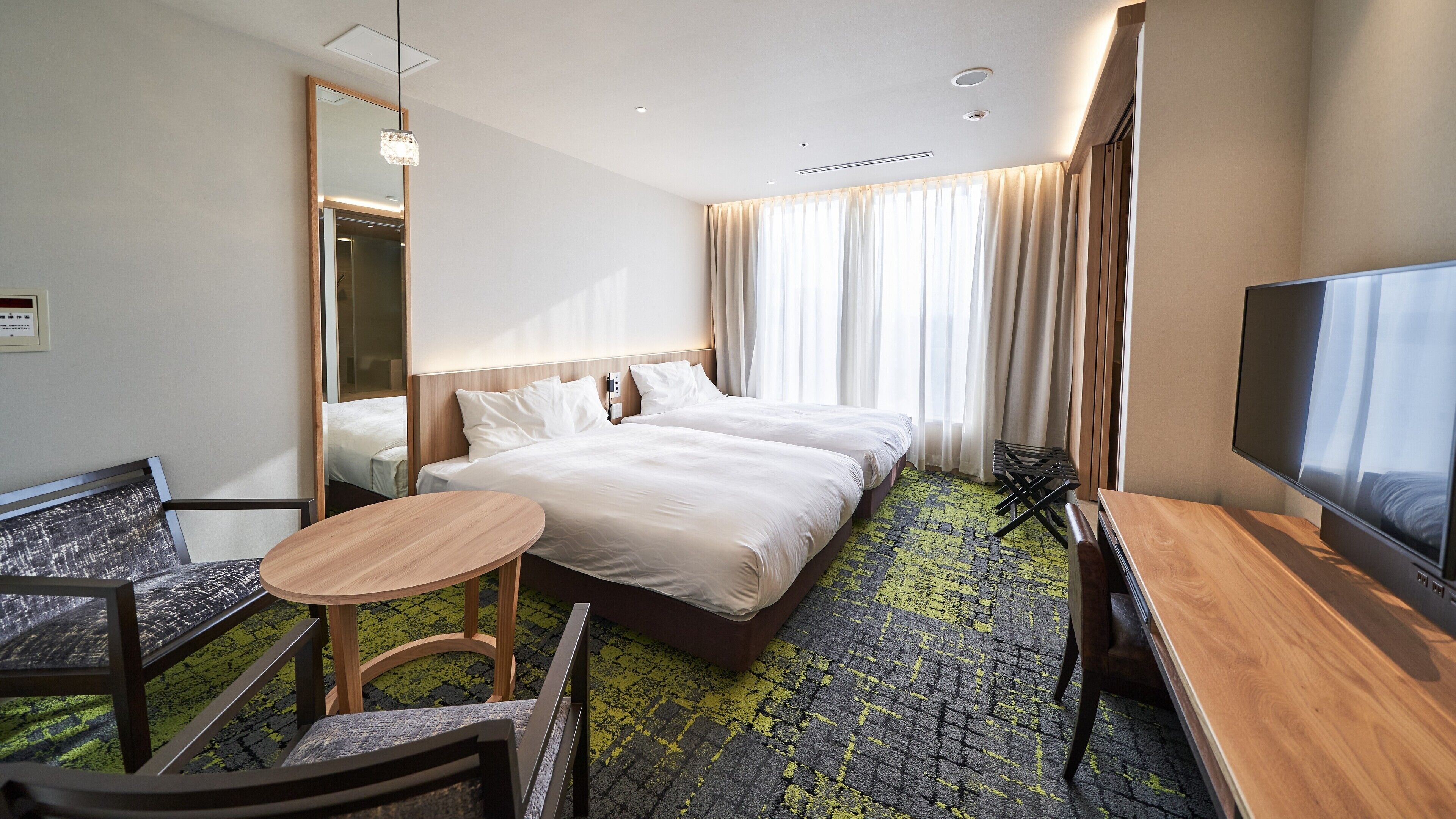 A hotel room with a bed, a TV, a desk, a chair, a small table, and a mirror.