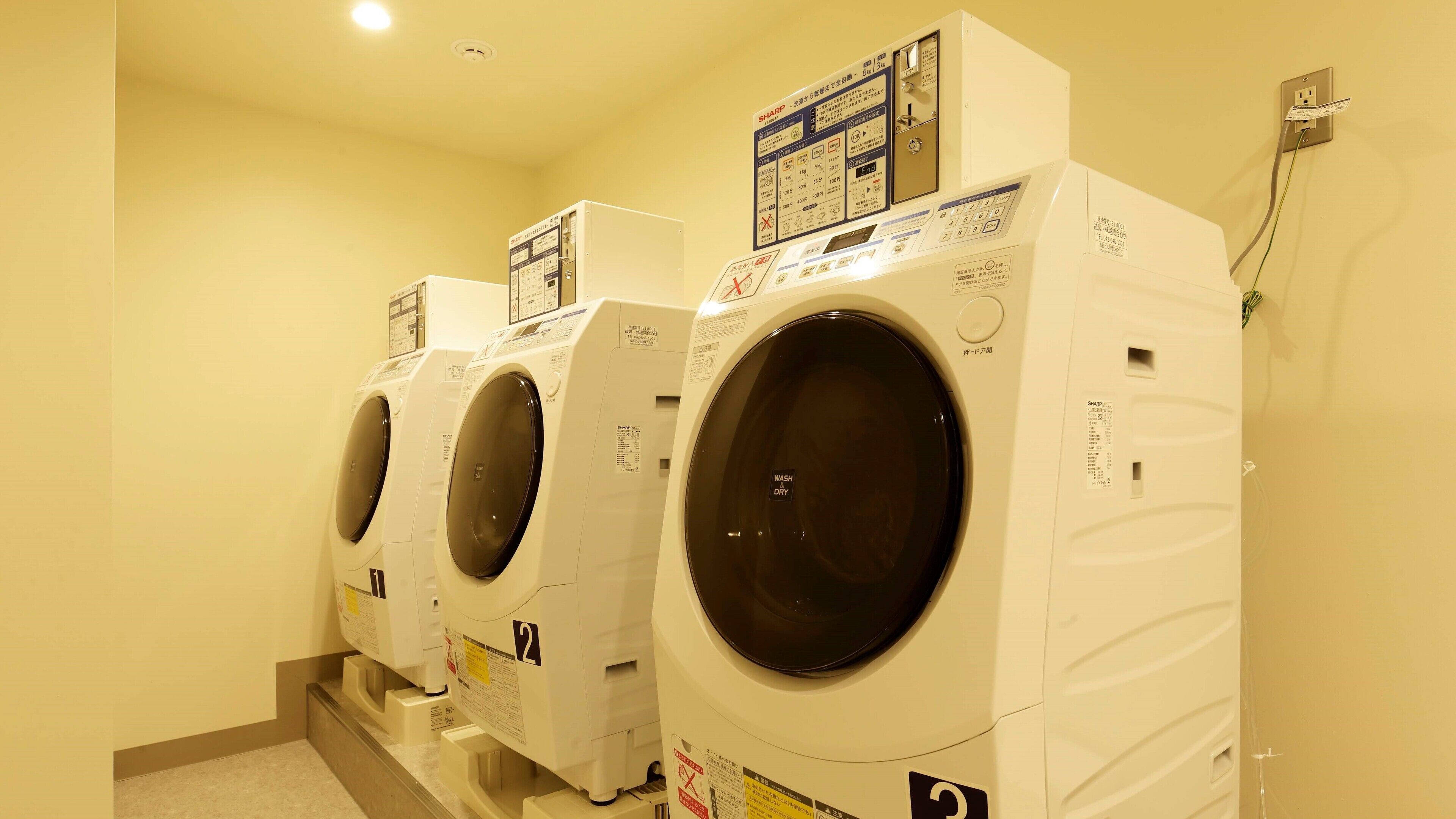 A row of white front-loading washing machines in a laundry room with a control panel above each machine.