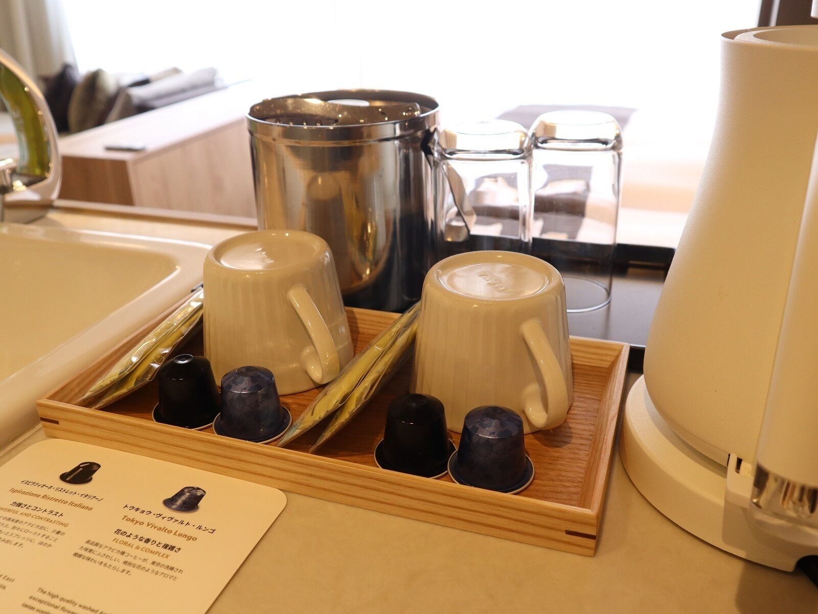 A tray with two white ceramic cups and three black cups, a stainless steel coffee pot, and a white electric kettle on a wooden tray.