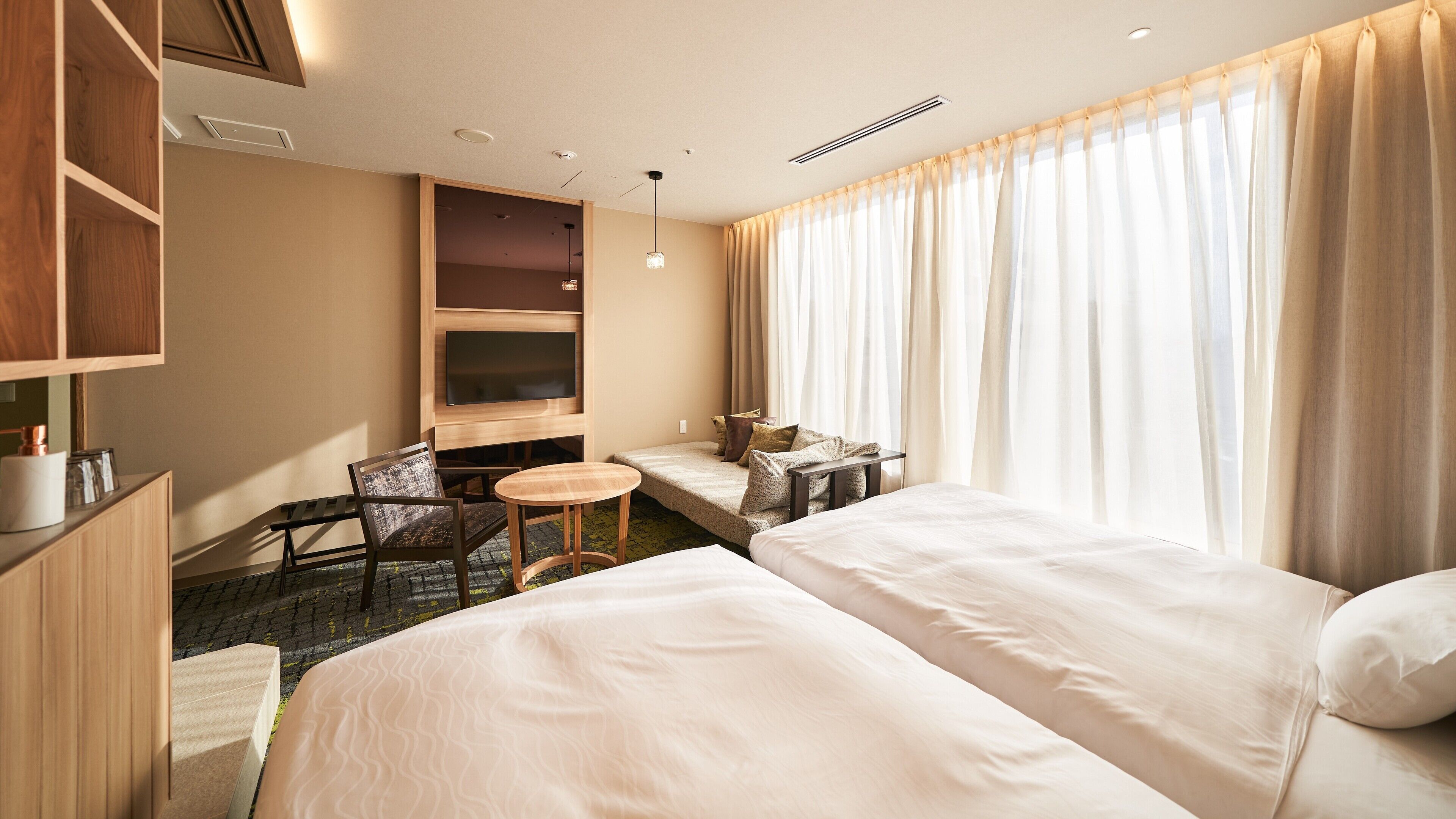 A hotel room with two beds, a small table, a chair, a TV, and a window with curtains.