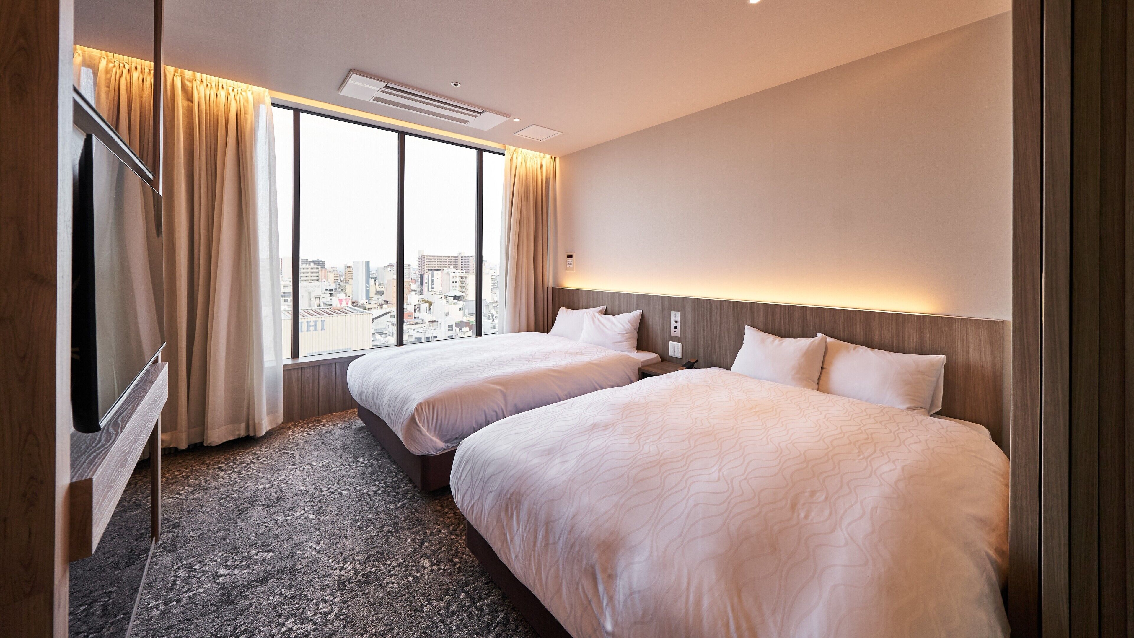 A hotel room with two beds, a TV, and a large window offering a city view.