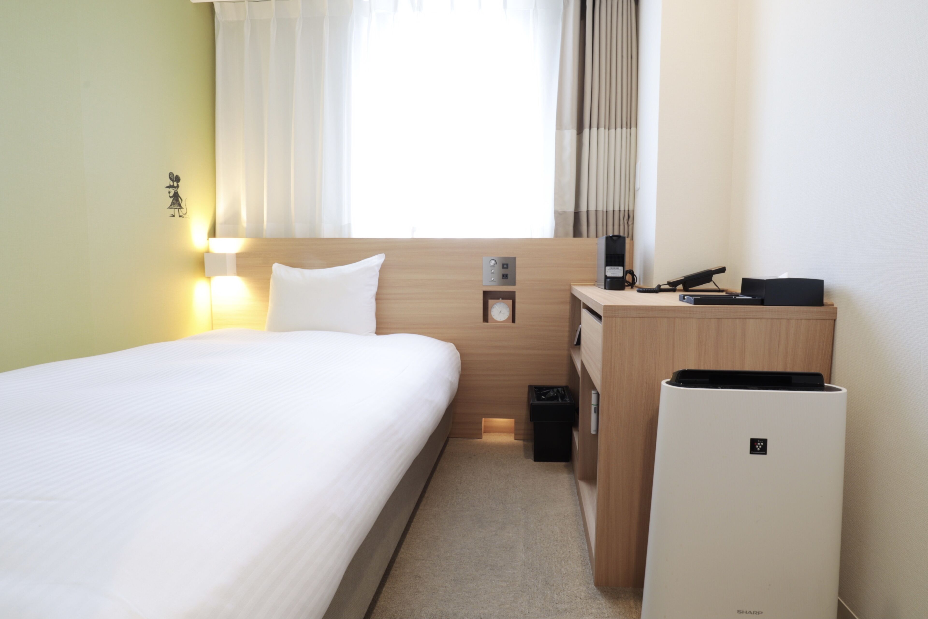A hotel room with a bed, bedside table, lamp, and a small desk with a phone and a coffee maker.