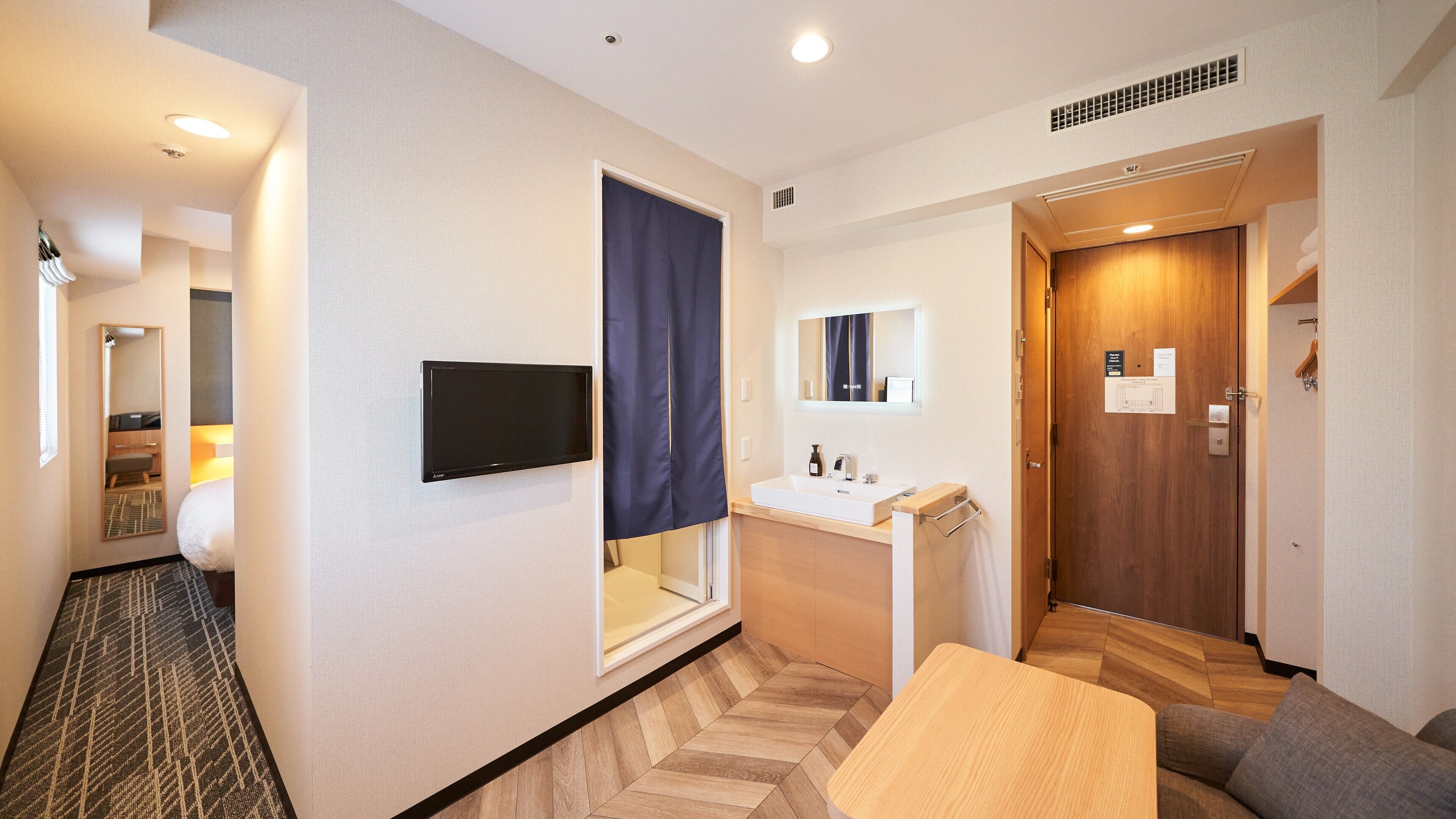 A hotel room with a flat-screen TV, a large window with a curtain, a sink, a mirror, a wooden door, and a small seating area.