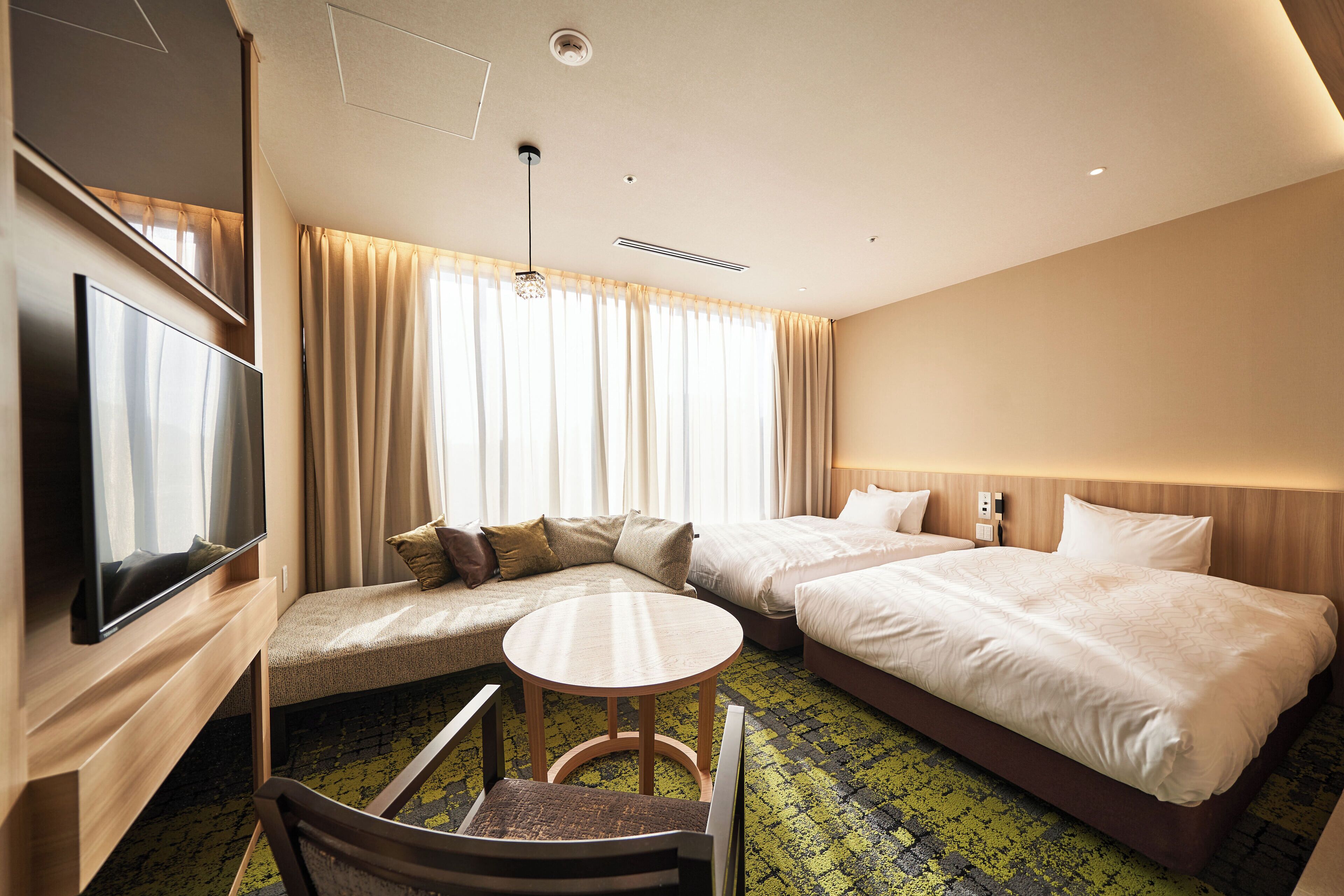 A hotel room with a TV, two beds, a sofa, a small table, and a chair.