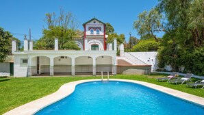 A swimming pool with a white building in the background, a covered patio area, and a fenced garden.