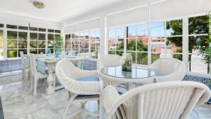 A sunlit room with a round glass table surrounded by wicker chairs, large windows offering a view of buildings and greenery.