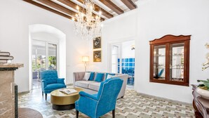 A spacious living area with a chandelier, blue armchairs, a sofa, and a coffee table. There is a fireplace, a dining area visible through an archway, and a large mirror on the wall.