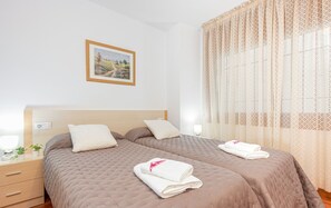 Uma cama de casal com duas almofadas, uma mesa de cabeceira com abajur, um quadro na parede e uma janela com cortinas leves.