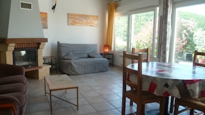 Een woonkamer met een open haard, een bank, een eettafel met stoelen en uitzicht op groen buiten het raam.