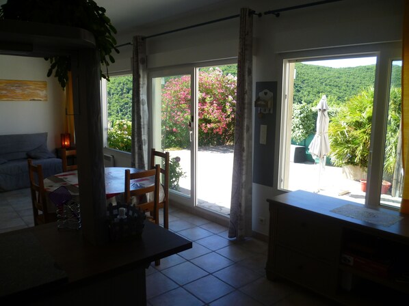 Una habitació amb taula de dinar, cadires, sofà i una gran porta corredissa de vidre que dona a una zona exterior amb vegetació i vistes a un turó.