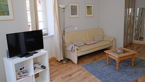 Una sala de estar con una televisión de pantalla plana sobre un soporte blanco, un sofá color beige, una mesa de centro de vidrio y una lámpara de pie.