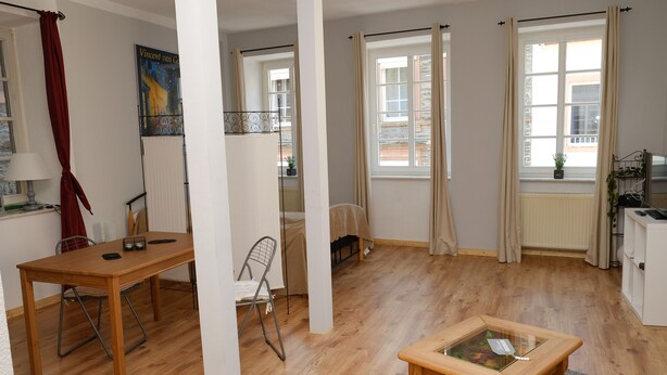 Una habitación con una mesa de madera, una silla, una mesa baja de vidrio y un ventanal con cortinas.