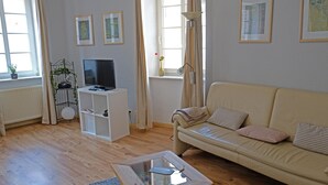 Una sala de estar con un sofá beige, una mesa de centro de vidrio, un televisor sobre un mueble blanco y una planta sobre un pedestal de metal.