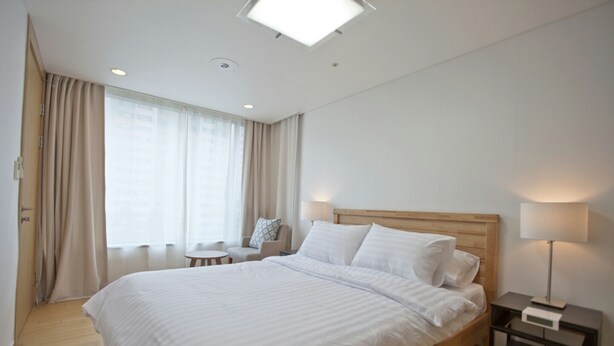 A modern hotel room with a large bed, a bedside table with a lamp, a small table, and a window with curtains.