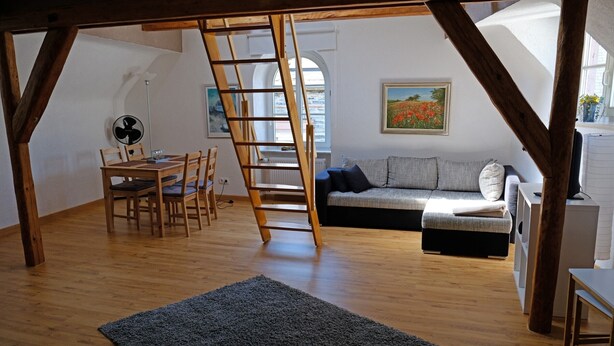 A loft-style room with a wooden floor, a gray sofa, a dining table with chairs, a ladder, and a fan.