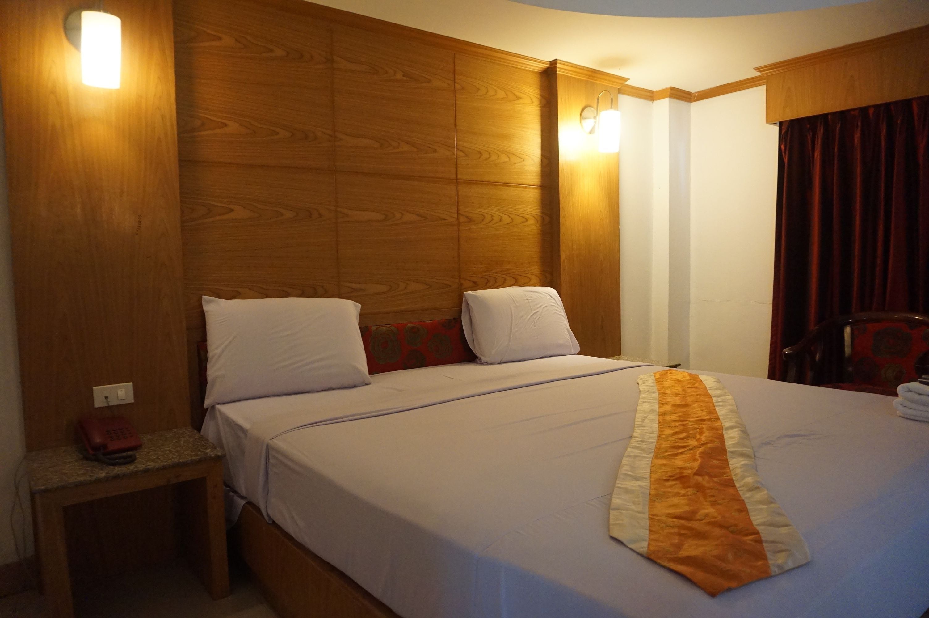 Une chambre d’hôtel avec une tête de lit en bois, un lit recouvert de draps blancs, une serviette à rayures rouges et blanches, et une petite table avec un téléphone.