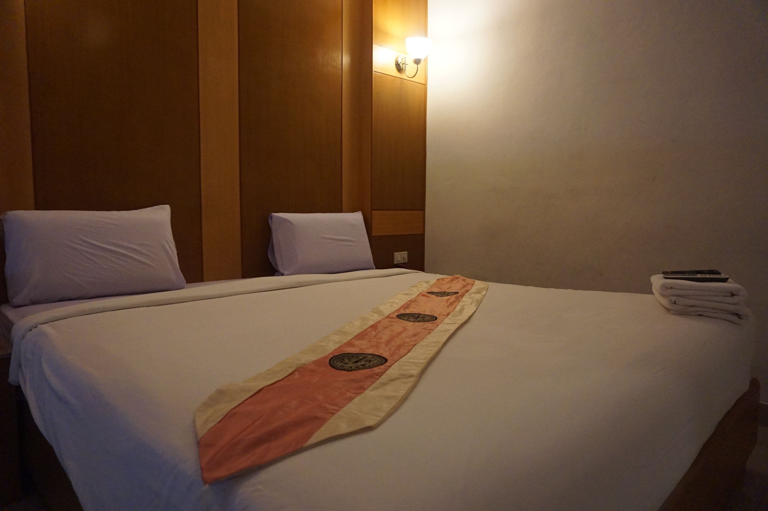 A hotel room with a large bed, a headboard, a wall-mounted light, and a folded towel on the bed.