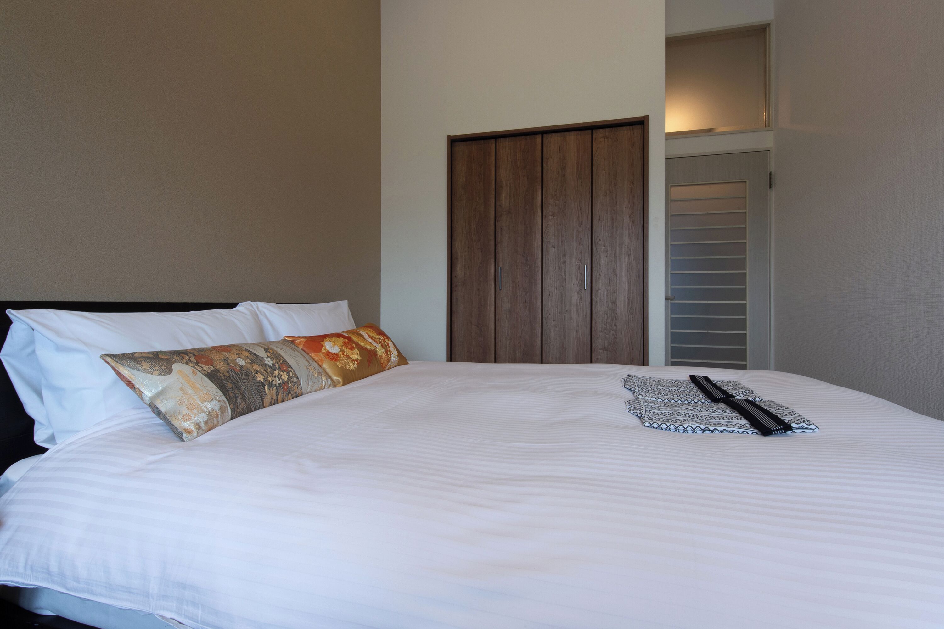 A neatly made bed with white linens and a patterned pillow, a wooden wardrobe, and a door with a window.