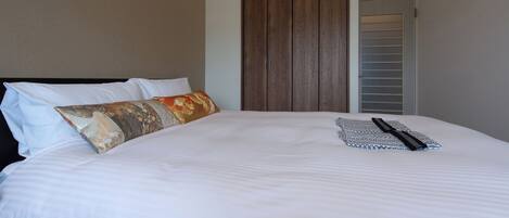 Una cama bien hecha con sábanas blancas y un cojín estampado, un armario de madera y una puerta con ventanal.