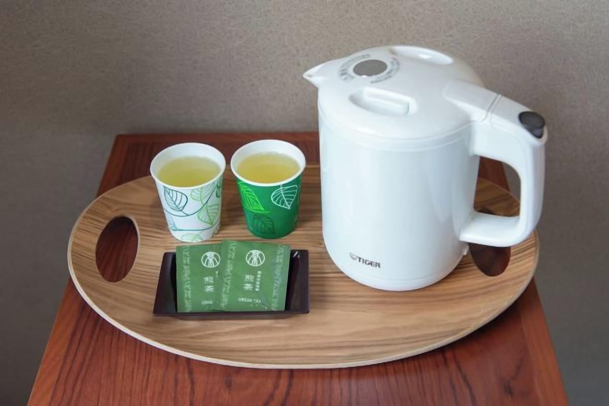Una tetera eléctrica blanca, dos vasos de papel con diseños de hojas, y una bandeja con una caja verde sobre una mesa de madera.