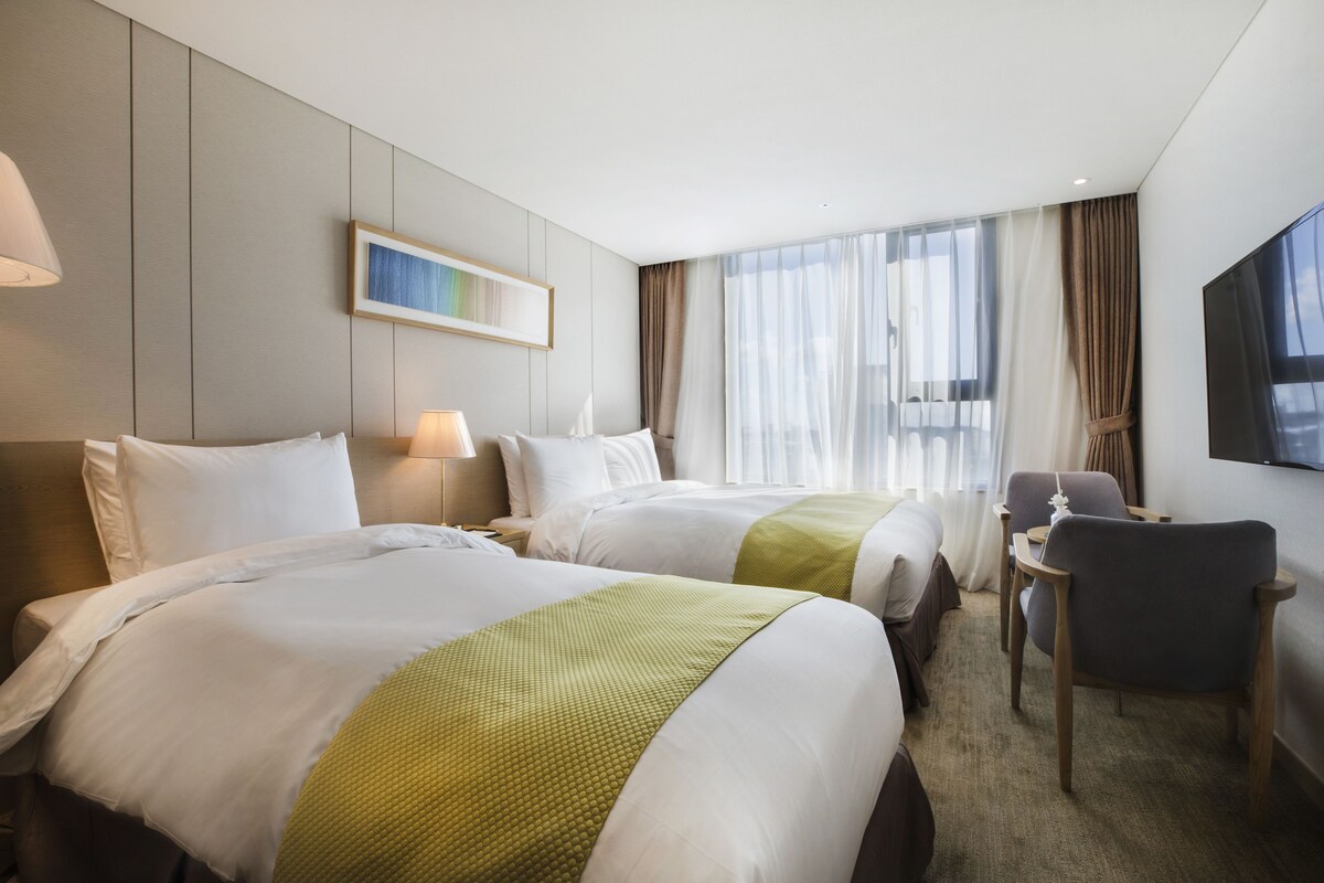A hotel room with two beds, a desk, a chair, a TV, and a window with curtains.