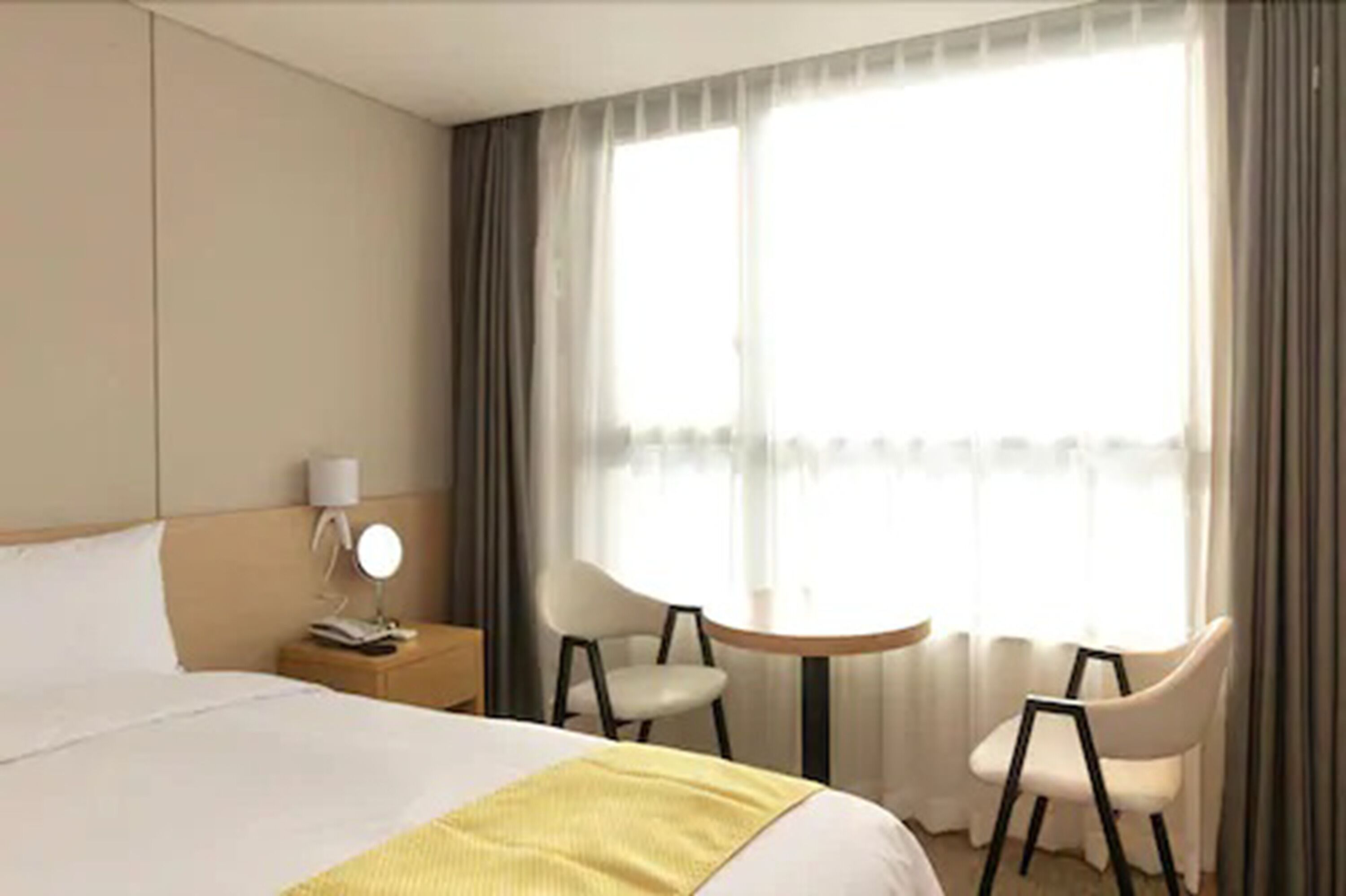 A hotel room with a bed, a bedside table with a lamp, two chairs, and a window with curtains.
