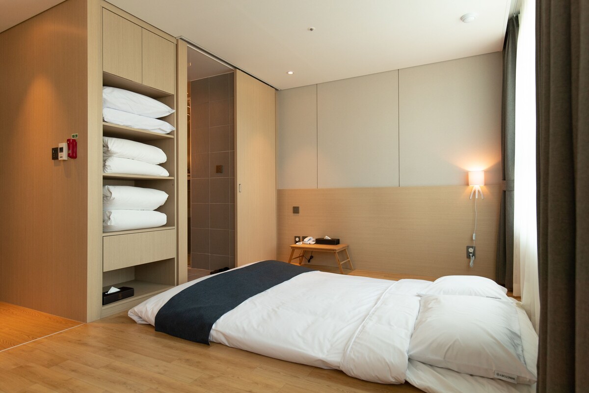 A hotel room with a bed, a wardrobe with hanging towels, a bedside table with a lamp, and a view of the outside.