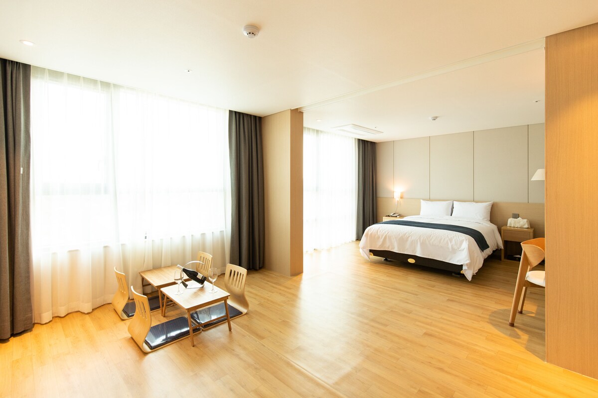 A hotel room with a bed, a desk with a chair, a table with a chair, and a window with curtains.