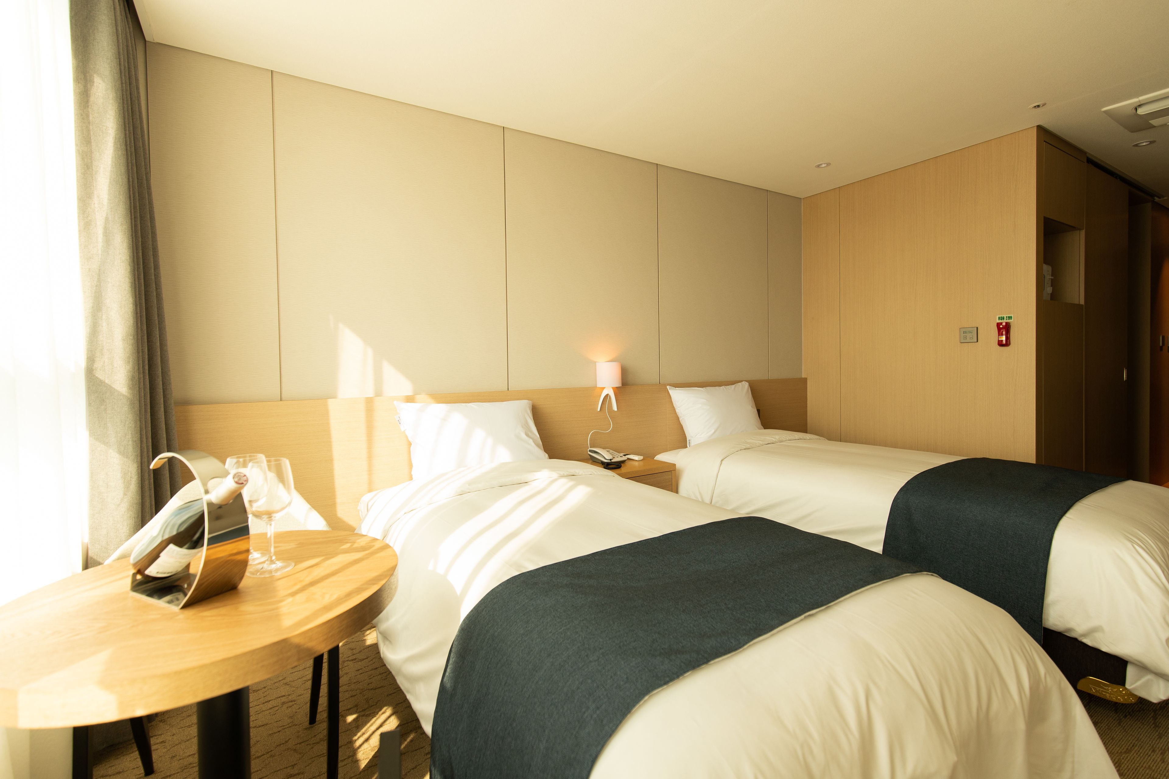A hotel room with two beds, a bedside table with a lamp, a telephone, and a window with curtains.