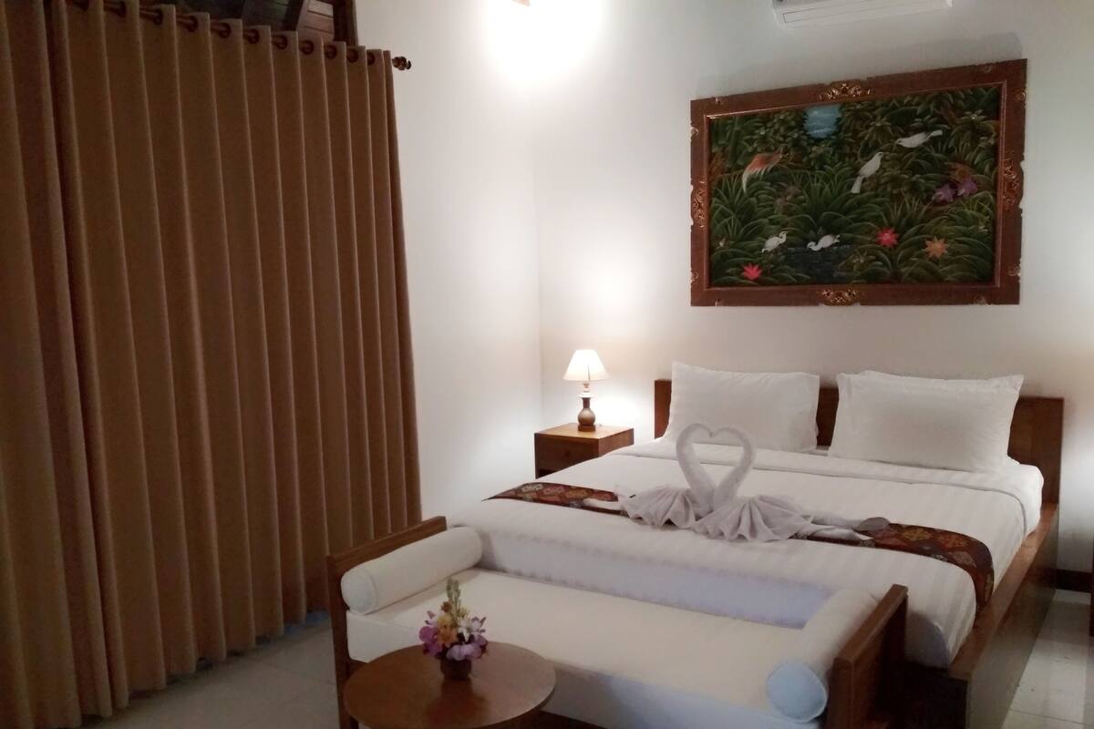 Una habitación de hotel con una cama, una mesita de noche, una lámpara, una pequeña mesa con una flor y un cuadro en la pared.