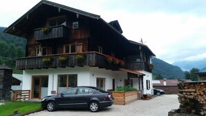 Ein zweistöckiges Holzhaus mit Balkonen, ein Auto davor und eine Bergkulisse im Hintergrund.