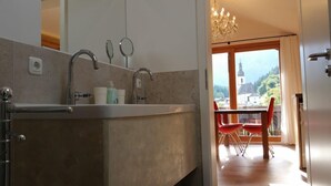 Ein modernes Badezimmer mit einem großen Waschbecken, zwei Wasserhähnen und einem Spiegel. Ein Fenster bietet Blick auf eine Kirche und Berge.