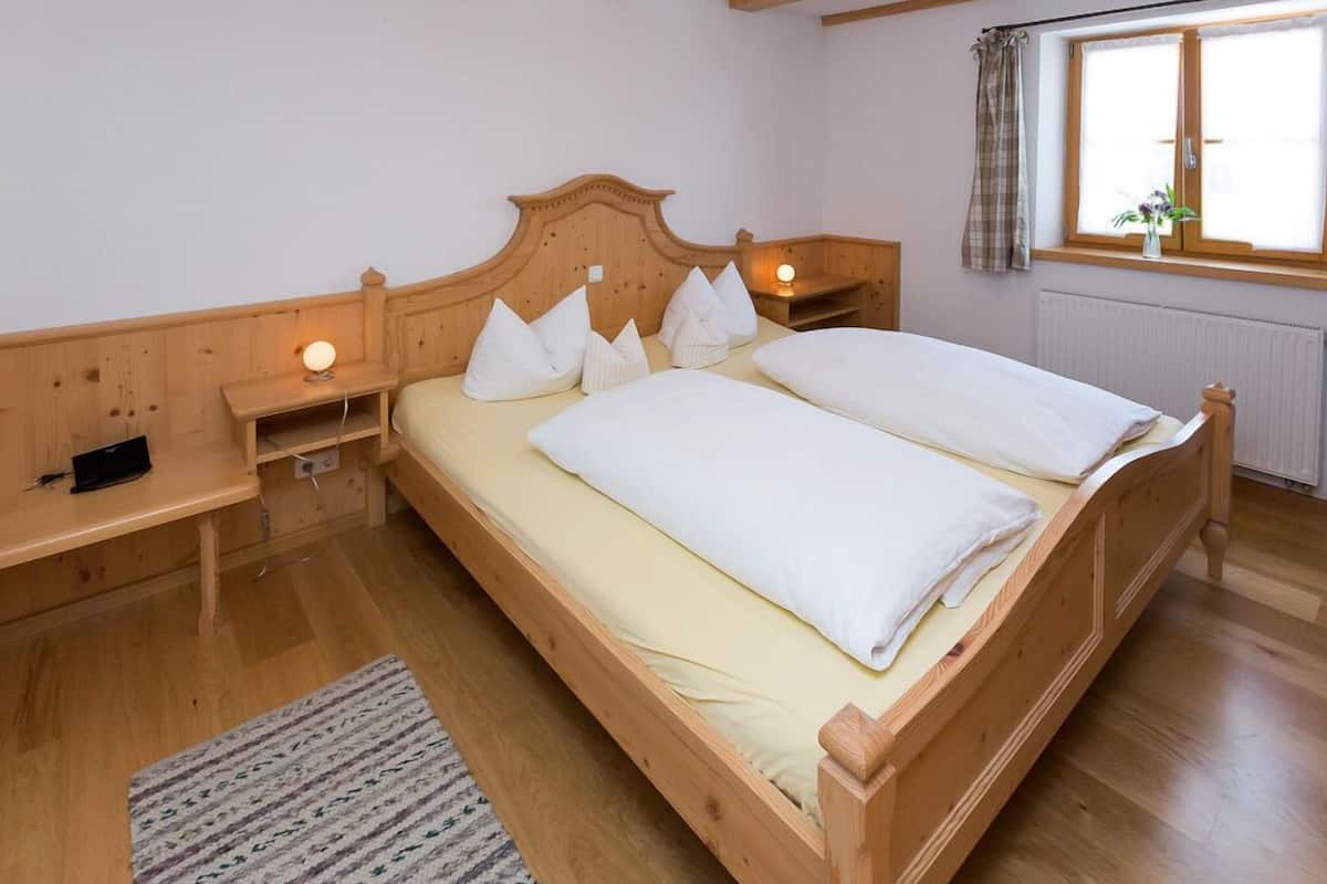 A wooden bed with white bedding in a room with wooden floors and a window with curtains.