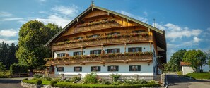 Un edificio de madera de varios pisos con balcones decorados con flores, un sendero de piedra y una pequeña estructura de madera al frente.