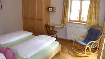 Una habitación con dos camas individuales, un armario de madera, una silla de mimbre, una mesita y una ventana con cortinas.
