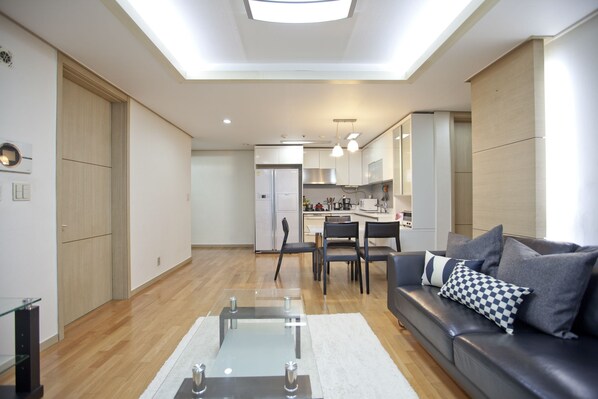 A modern living room with a glass coffee table, a sofa with patterned cushions, and a kitchen in the background.