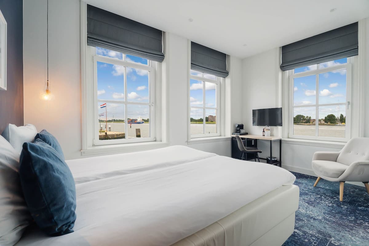 A hotel room with a large bed, a desk with a computer, a TV, and a chair. There are large windows with views of the outside.
