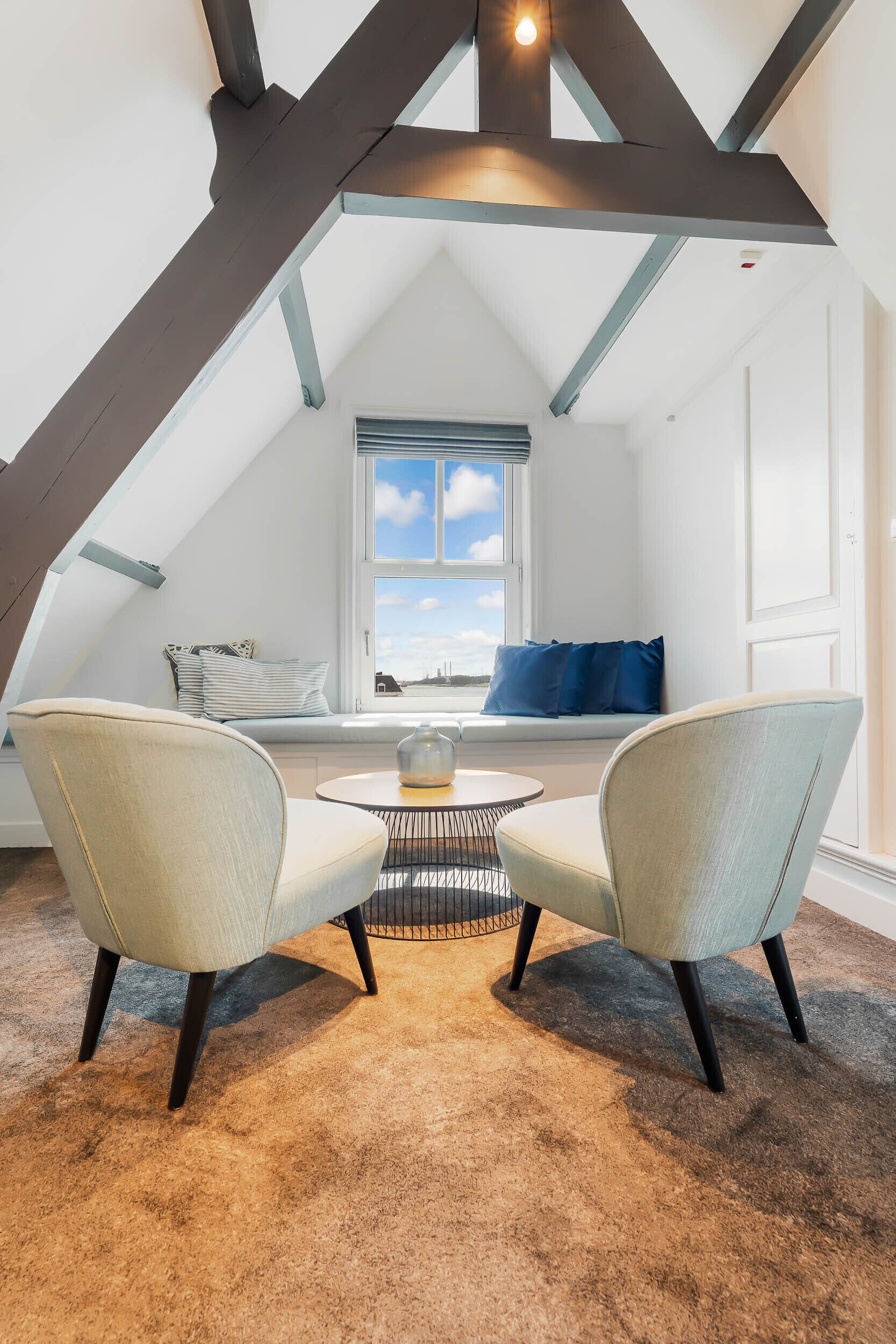 Een gezellige zolderkamer met schuine plafonds, een ronde tafel, twee fauteuils, een raam met uitzicht en een ingebouwde bank met kussens.