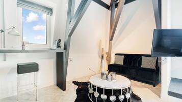 A modern room with a round drum table, a black leather sofa, a TV, and a window with a view of the sky.