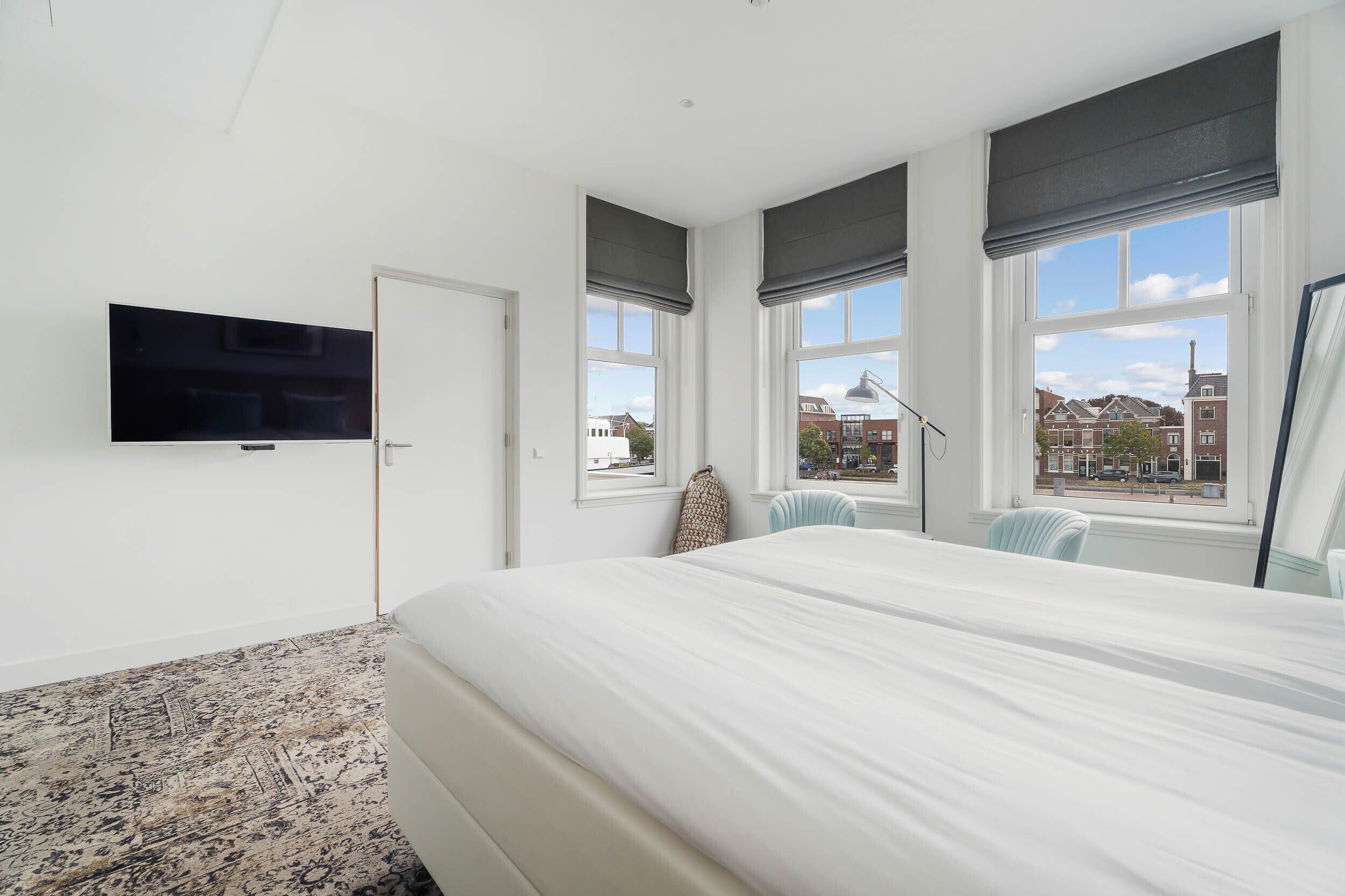 A hotel room with a large bed, a flat-screen TV mounted on the wall, and large windows with dark curtains.