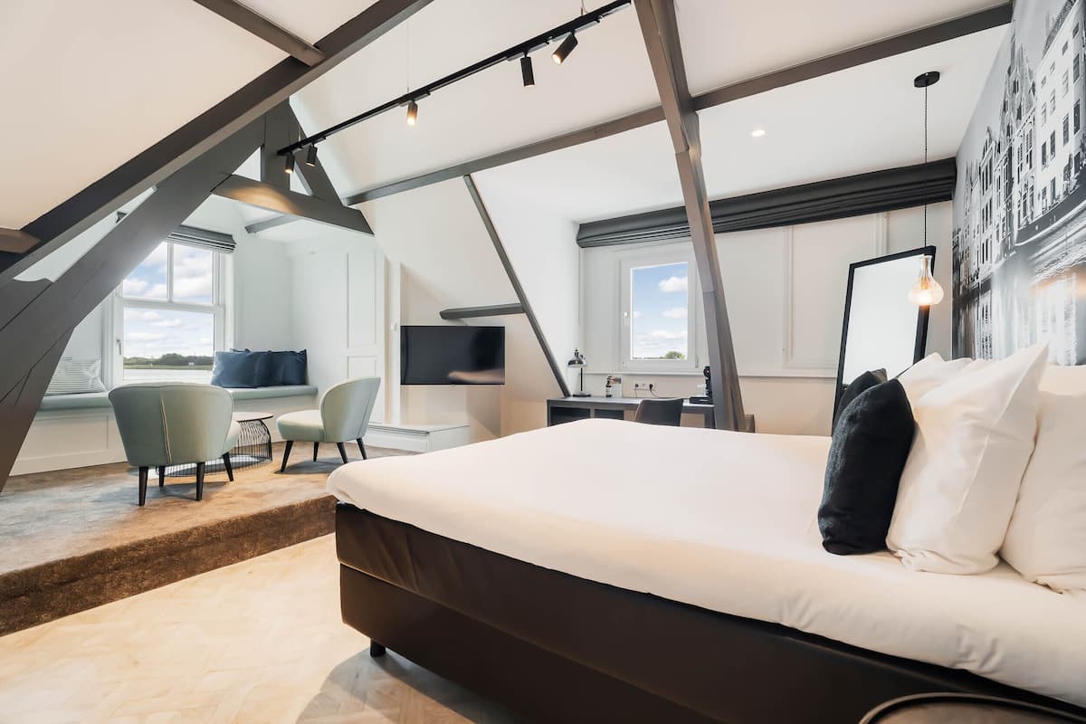 A modern bedroom with a large bed, a desk, and a chair. There is a window with a view of the sky and a cityscape.