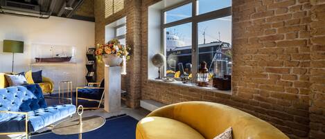 A modern living room with a yellow sofa, a blue sofa, and a round table. There is a large window with a view of a ship and a brick wall.