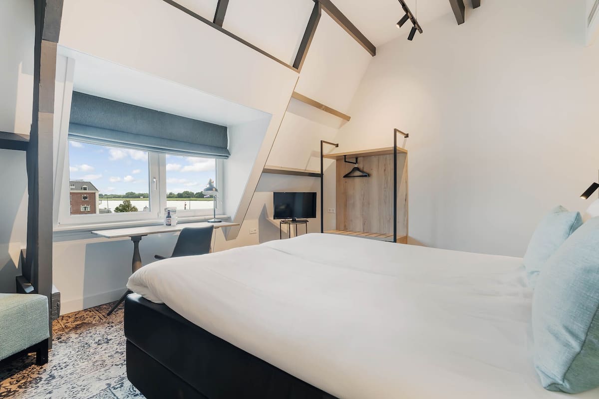 A bedroom with a bed, a desk, a chair, a TV, and a window with a view of a building and trees.